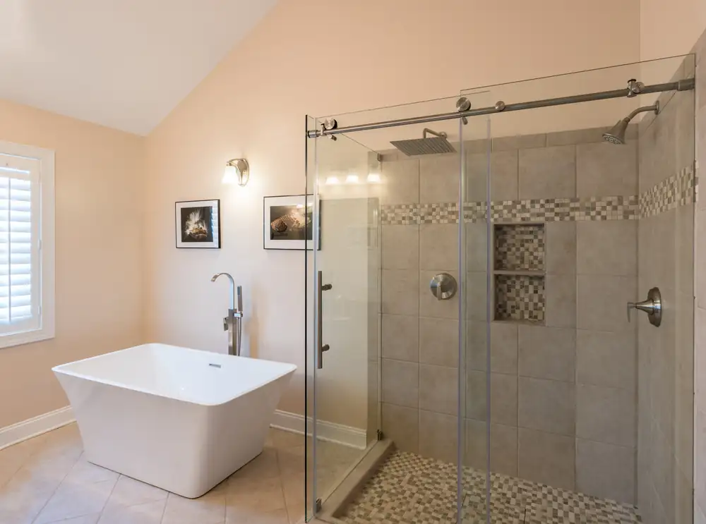 Bathroom remodeling Warren MI: frameless sliding glass shower door on a tiled walk-in shower with a mosaic niche, beside a freestanding soaking tub