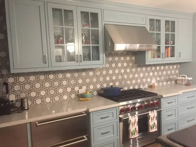 Kitchen with pale blue glass-front cabinets, a hexagon mosaic backsplash, quartz counters and a stainless range hood